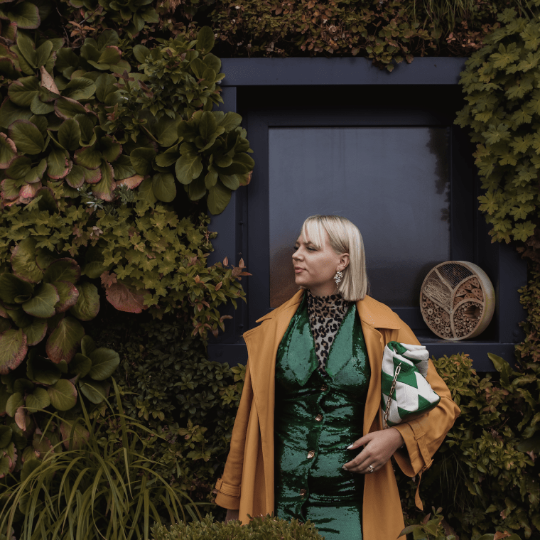 Cassandra Dittmer, a woman with blond short hair and fashionable clothing, stands in front of a lush green space.